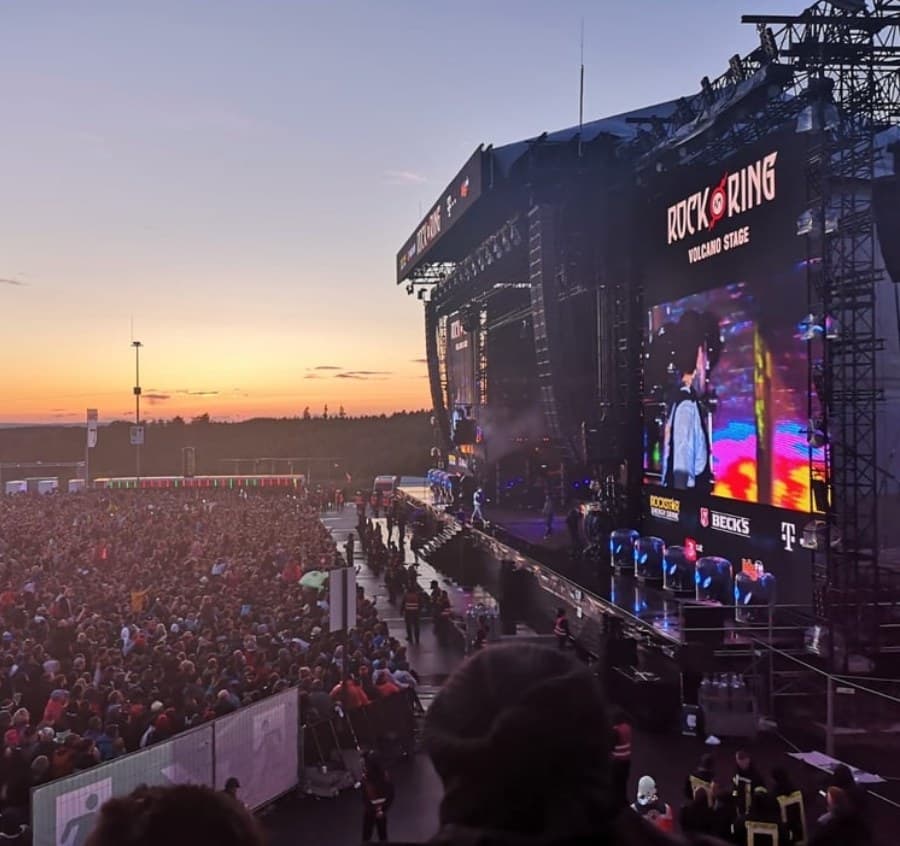 Rock am Ring Festivalbühne bei Sonnenuntergang, mit "ROCK AM RING VOLCANO STAGE" auf großer Leinwand und Menschenmenge.