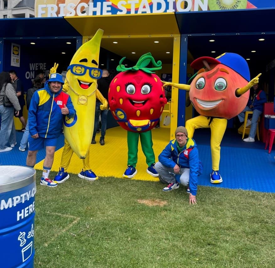 Drei Personen in Obst-Maskottchen-Kostümen mit zwei Männern vor einem blauen und gelben "FRISCHE STADION"-Stand.